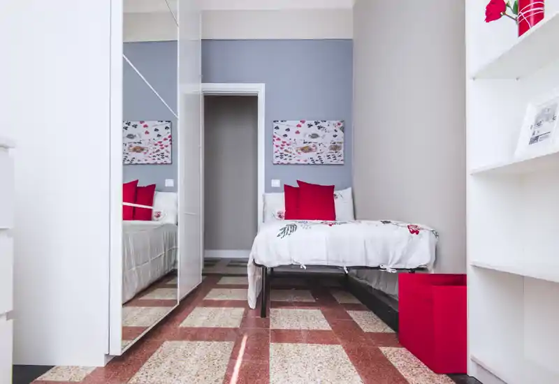 Bright, modern single bedroom with a neatly made bed, red accent cushions and mirrored wardrobe that enlarges the space. Clean lines and tasteful decor make it suitable for short-term rentals or a cozy private room.