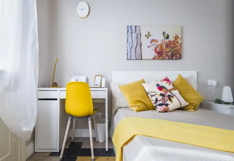 Bright, stylish bedroom featuring a comfortable bed with decorative cushions, a small desk with a yellow chair, and tasteful wall art — ideal for a cozy single or couple stay.