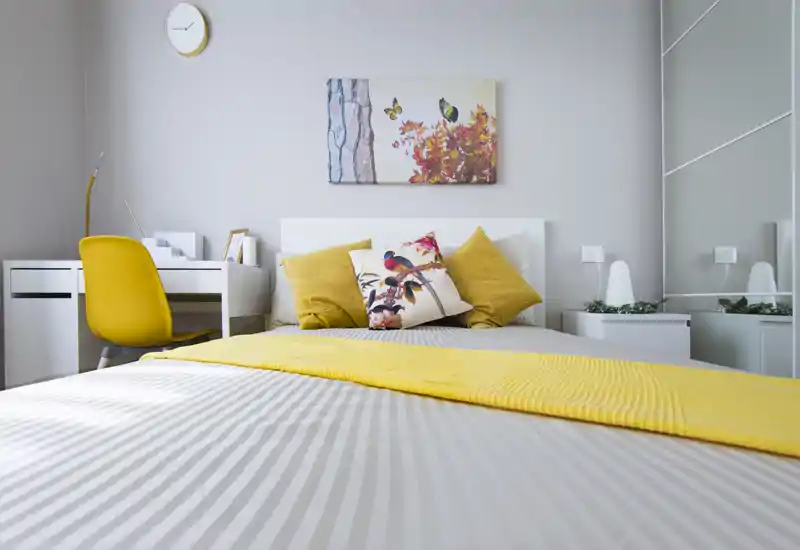 Bright, stylish bedroom with a large bed dressed in striped linens and a yellow runner, decorative cushions and a small desk with a chair — ideal for a cozy rental listing.