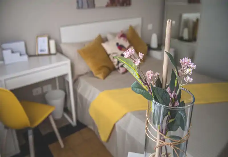 Dormitorio acogedor y bien presentado con ropa de cama beige y toques mostaza; cojines decorativos y una manta brillante crean un ambiente cálido.