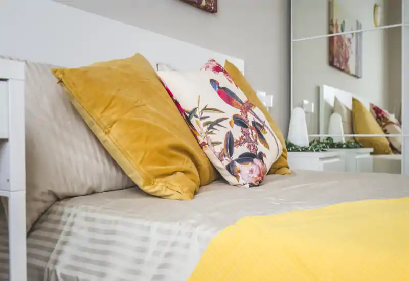 Bright, cozy bedroom close-up with decorative cushions and a neatly made bed featuring warm yellow accents and a patterned throw pillow.