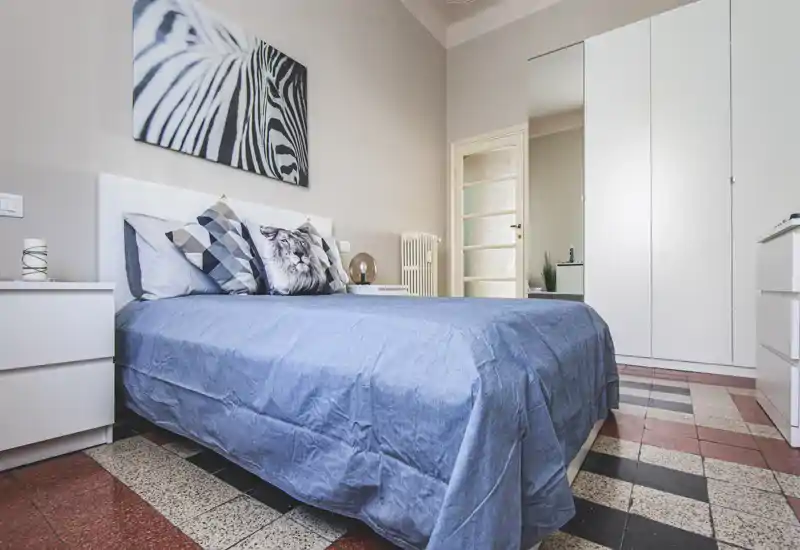 Bright, neatly staged bedroom with a full visible double bed, patterned cushions and a large wardrobe; calm neutral tones and tiled floor create a clean, welcoming space.