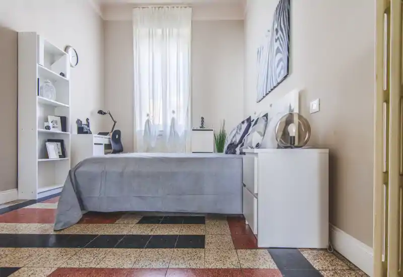 Bright, neatly styled bedroom with a full bed, bedside table and shelving; large window provides abundant natural light and tasteful minimal decor.