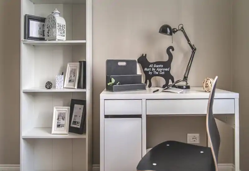 Coin bureau compact soigné avec un bureau blanc, des étagères et une lampe de bureau noire. Des éléments décoratifs — photos encadrées, petite plante et un panneau en forme de chat — apportent une touche chaleureuse.