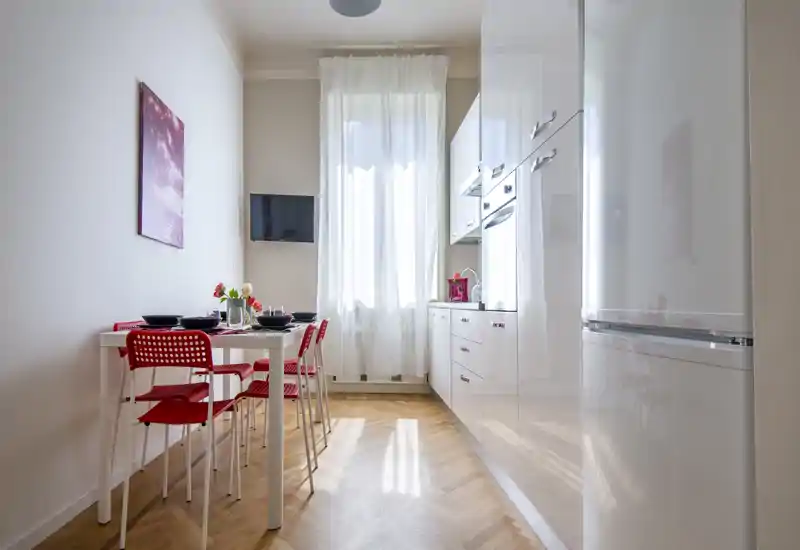 Cuisine moderne et lumineuse avec meubles blancs, petite table à manger et chaises rouges ; la grande fenêtre apporte une lumière naturelle et une ambiance aérée.
