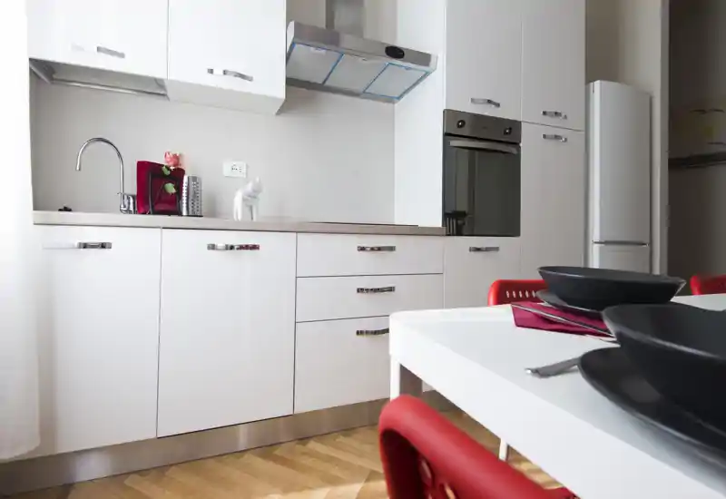 Cuisine moderne équipée avec placards blancs laqués, four et réfrigérateur encastrés, table blanche avec chaises rouges et vaisselle noire minimaliste.