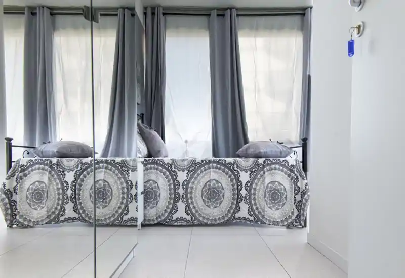 Bright, minimalist bedroom with a patterned bedspread, decorative pillows and soft gray curtains; mirrored wardrobe doors reflect the room and increase perceived space.