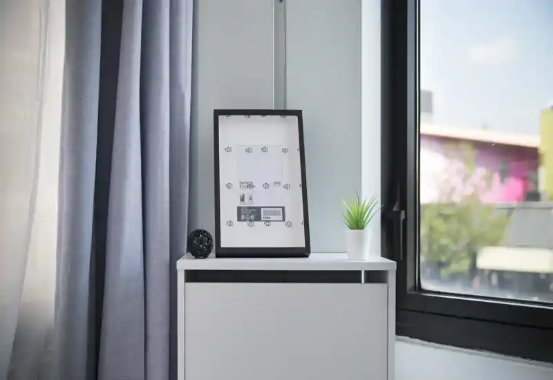 Bright, minimal bedroom corner featuring a white bedside cabinet topped with a framed print mockup, small decor object and a potted plant beside a large window with grey curtains.