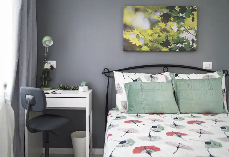 Bright, styled bedroom with a metal headboard, patterned duvet and green cushions. A small desk and chair sit by the window, creating a functional sleeping and work corner.