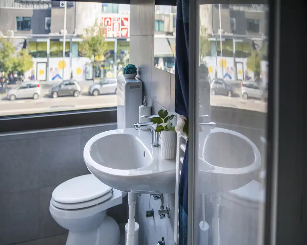 Salle de bain compacte et moderne avec lavabo blanc, toilettes et une grande fenêtre donnant sur la rue ; une petite plante et un distributeur de savon apportent une touche soignée.