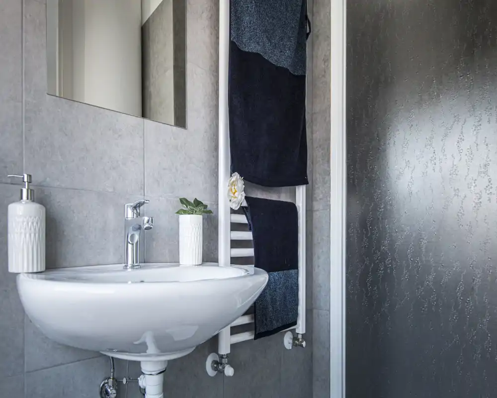 Salle de bain moderne et compacte avec lavabo mural, robinet chromé, miroir et porte-serviettes chauffante avec serviettes, à côté d'une porte de douche en verre texturé.
