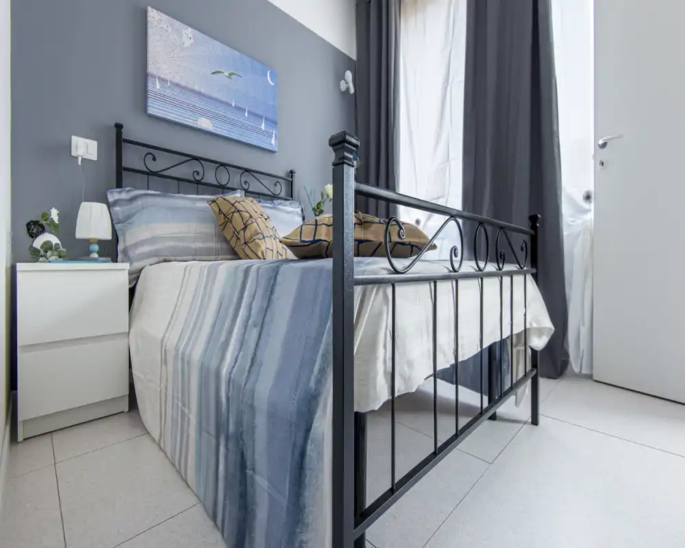 Chambre lumineuse et bien mise en scène avec lit en métal, literie rayée et table de chevet avec lampe ; mur gris et rideaux neutres pour une ambiance apaisante.