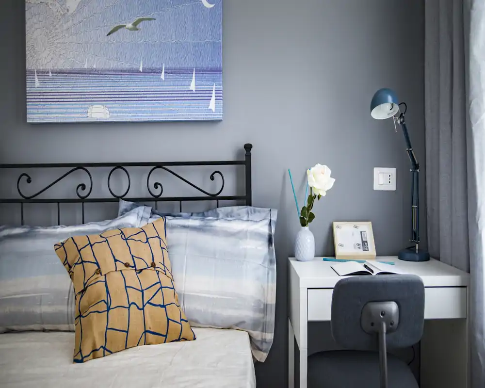 Coin chambre soigné et lumineux avec tête de lit en métal, coussin graphique et petit bureau avec lampe — agréable et accueillant pour une personne.
