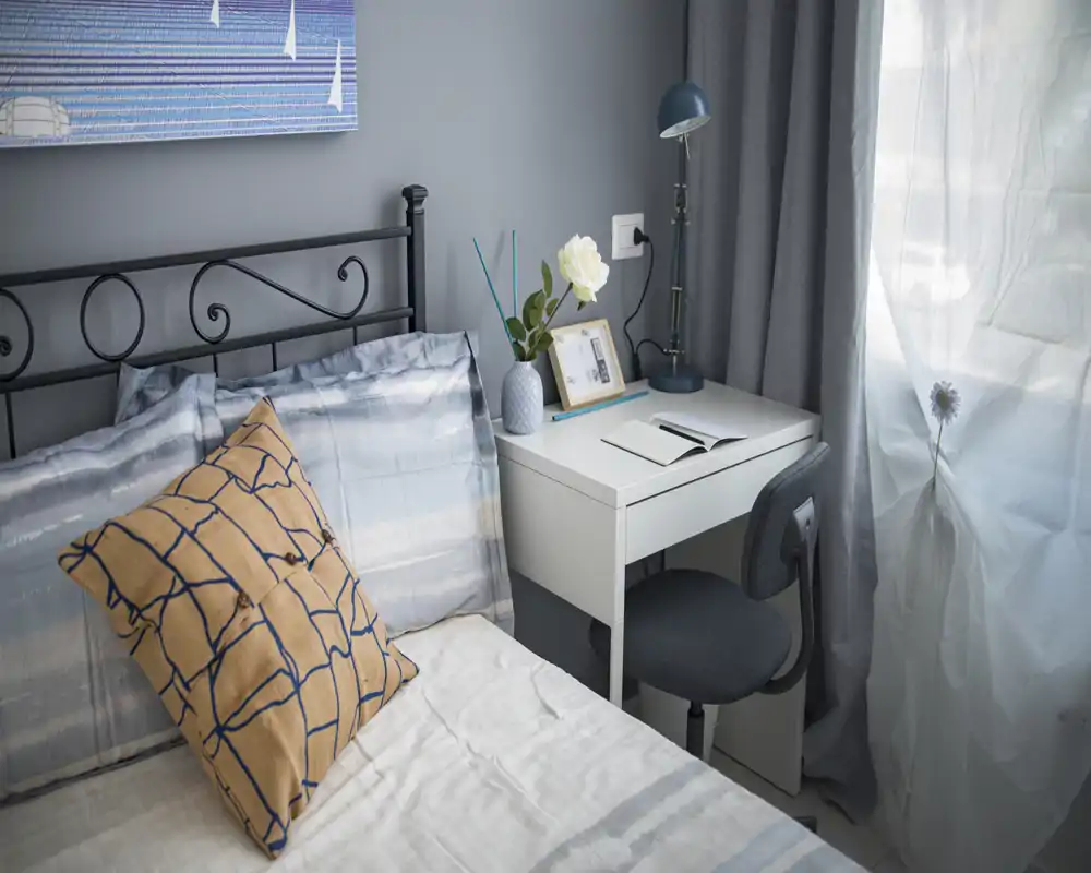 Coin de chambre lumineux et soigné avec coussin décoratif, tête de lit en fer et une petite table de chevet blanche avec lampe — parfait pour une chambre individuelle chaleureuse.