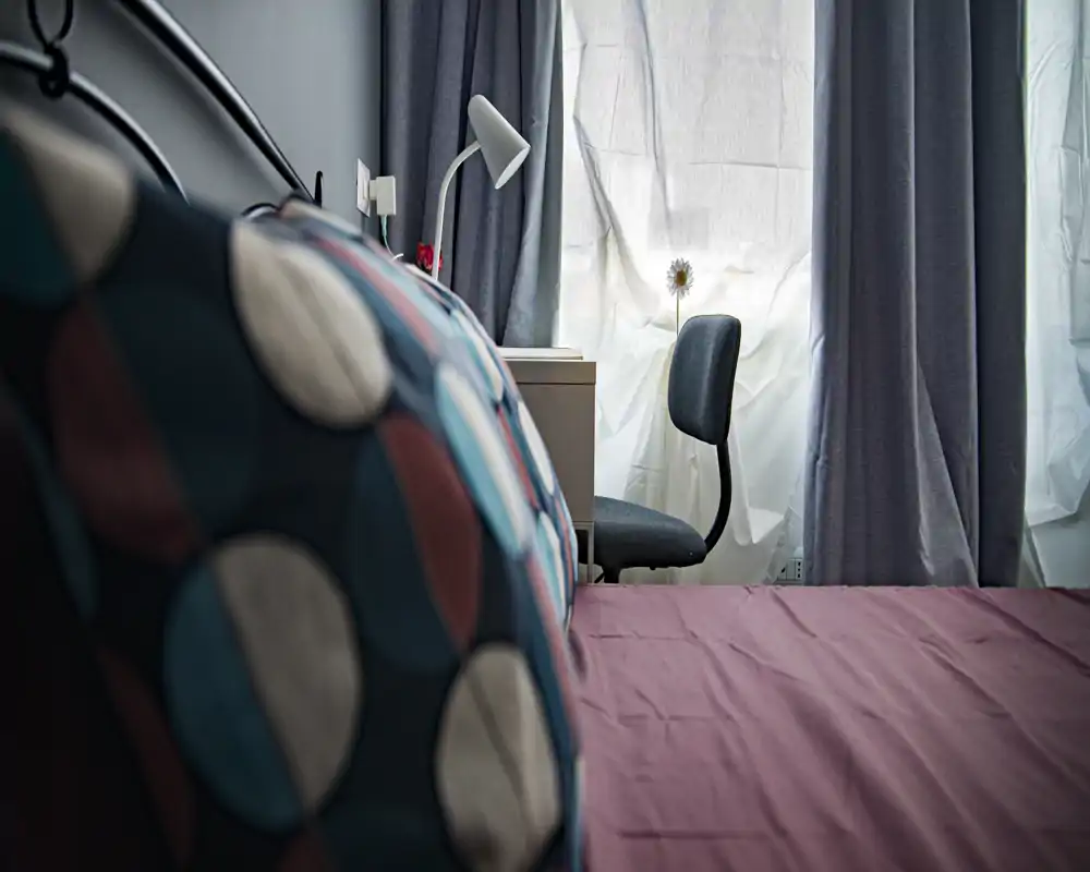 Bright, cozy bedroom corner with a patterned cushion in the foreground, a neatly made bed and a small desk with a lamp by the window providing soft natural light.