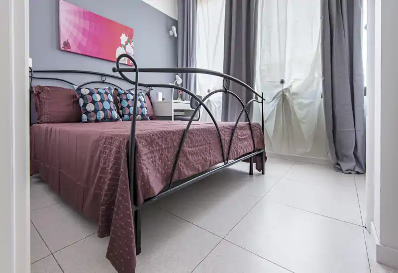 Bright, modern bedroom with a decorative metal bedframe, patterned cushions and coordinated bedding. Large window with curtains provides soft natural light and a neat bedside table completes the layout.
