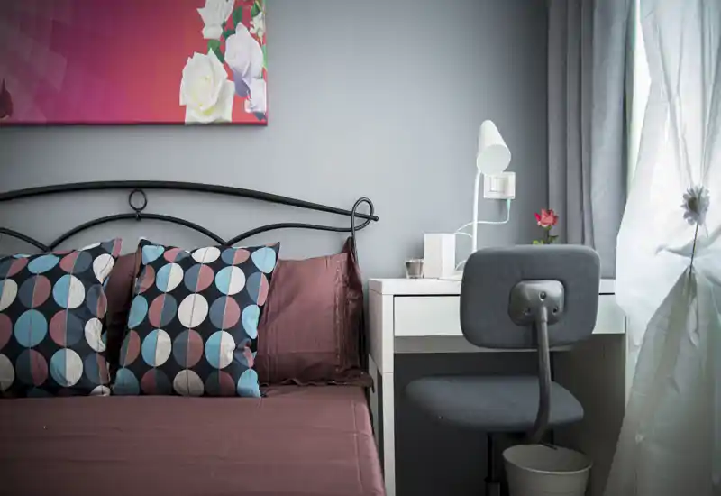 Bright, stylish bedroom corner with a patterned-pillows bed, bedside desk and lamp next to a window—cozy and well lit.