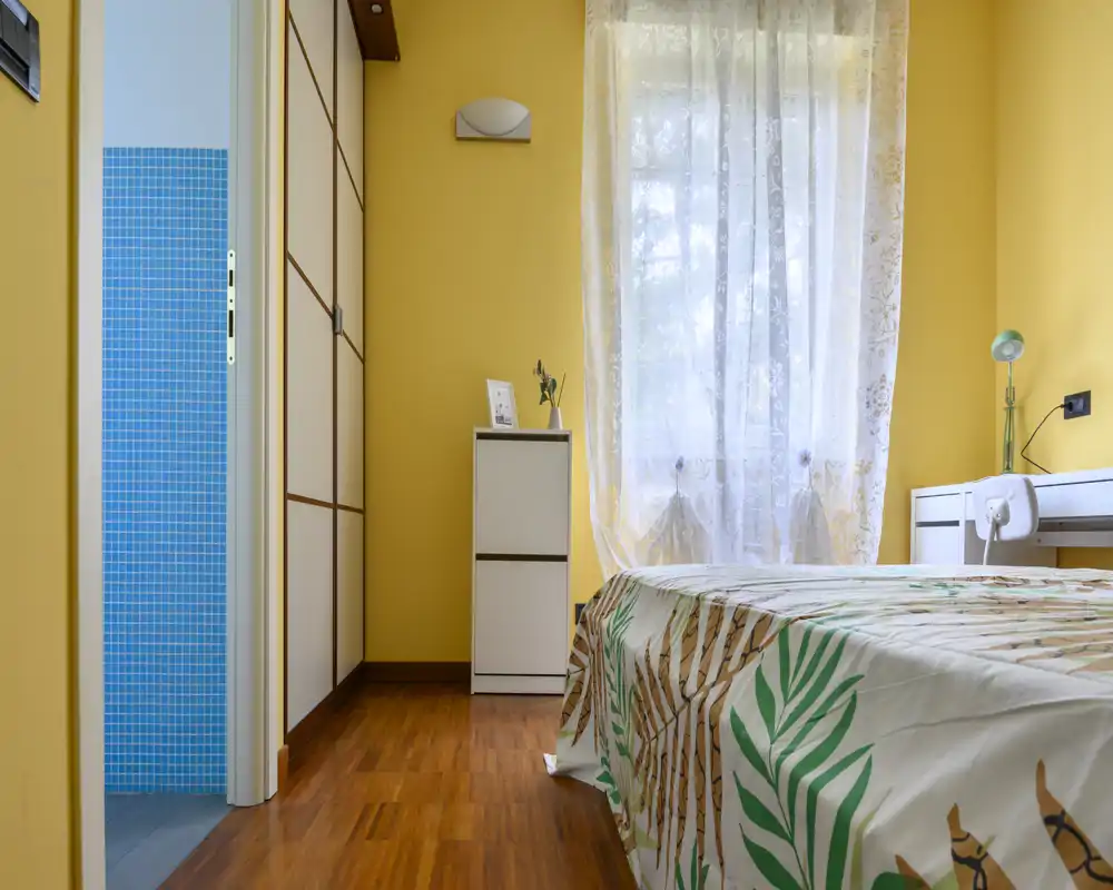 Dormitorio luminoso y ordenado con colcha estampada, escritorio blanco y armario. La luz natural suave atraviesa las cortinas de encaje, creando un ambiente cálido y acogedor.