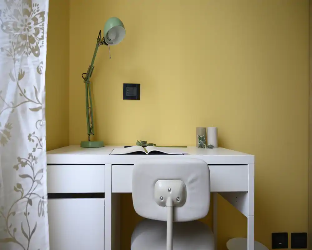 Espacio de trabajo compacto con escritorio blanco, silla acolchada y lámpara verde frente a una pared amarilla cálida — un rincón ordenado y bien iluminado perfecto para trabajar concentrado.