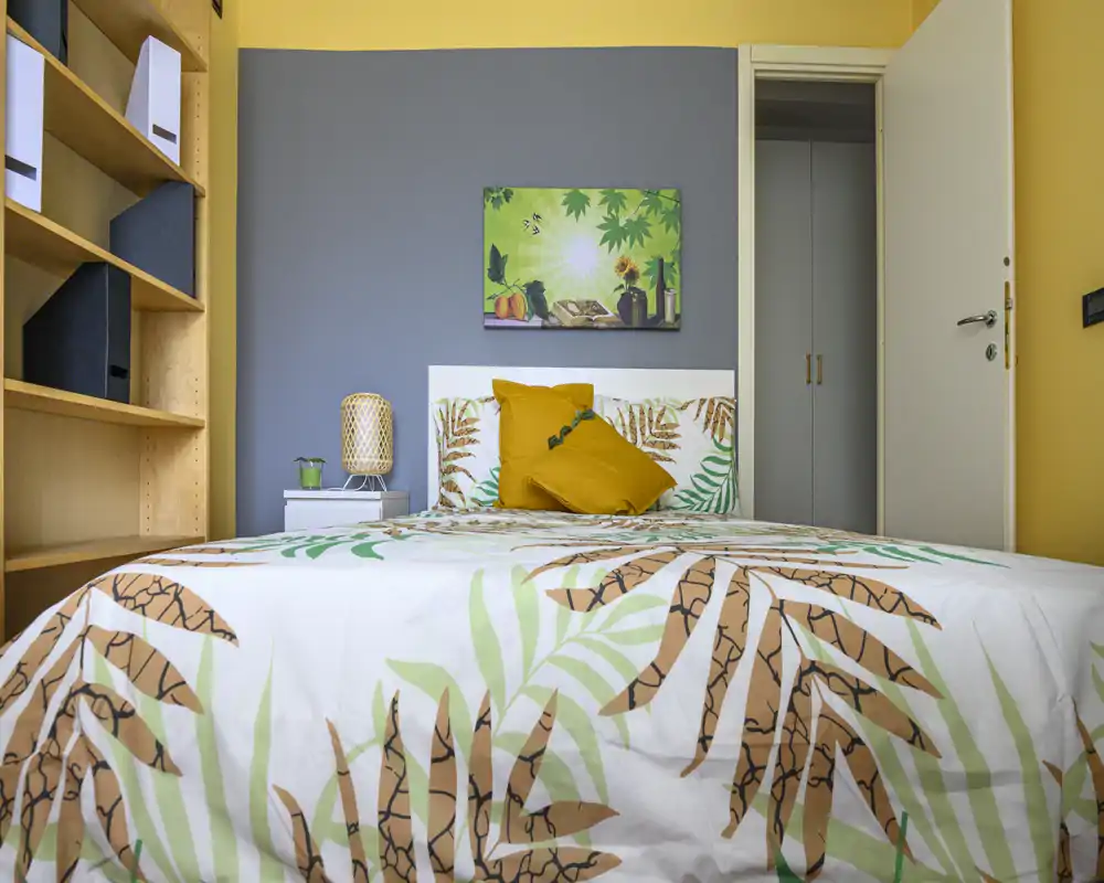 Dormitorio luminoso y bien decorado con colcha estampada, cojines decorativos y una lámpara de noche; estanterías neutras y pared en color de acento completan el ambiente.