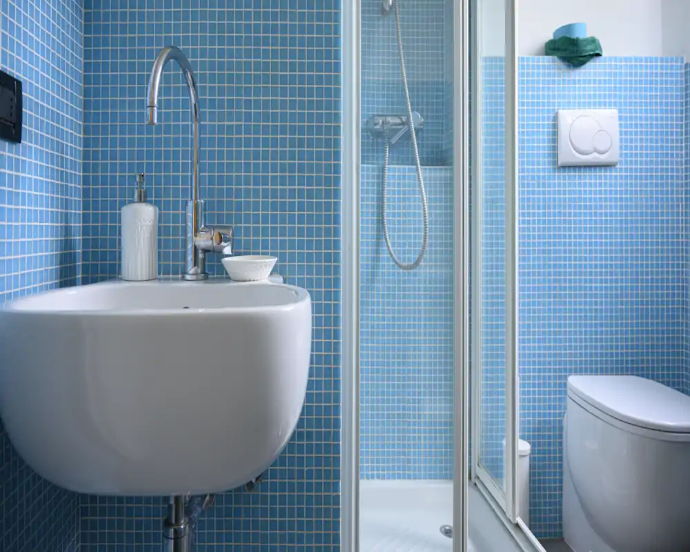 Baño moderno y compacto con azulejos tipo mosaico azules, lavabo mural, ducha con mampara de cristal y WC. Instalaciones limpias, luminosas y bien cuidadas.