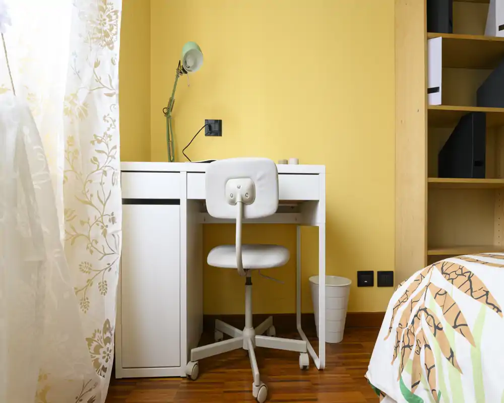 Rincón de dormitorio acogedor con escritorio y silla blancos junto a la cama. Pared amarilla y luz natural desde la ventana con cortina que crean un ambiente agradable.