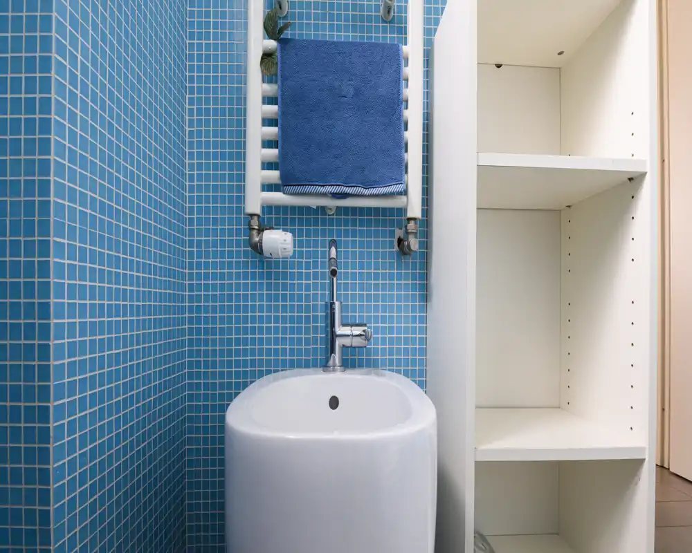 Rincón de baño moderno con azulejos de mosaico azul, un bidé/pequeño lavabo con grifo cromado y un toallero calefactado con una toalla azul — limpio y bien iluminado.