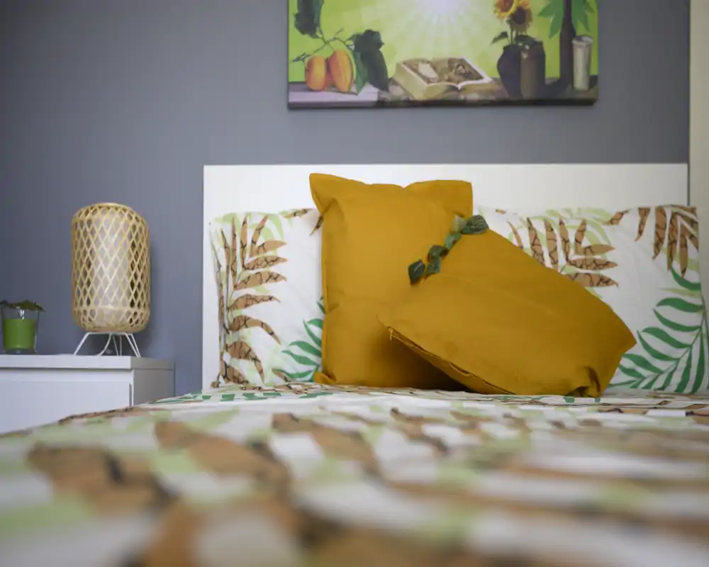 Dormitorio luminoso y con estilo: colcha estampada y dos cojines decorativos color mostaza sobre un cabecero blanco; la luz suave resalta la paleta de colores y la lámpara de mesita.