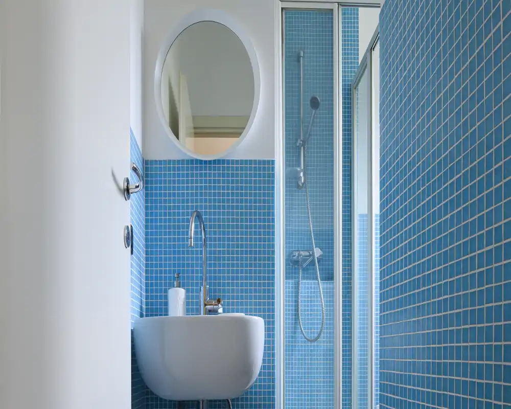 Baño moderno y compacto con azulejos mosaico azules, lavabo suspendido, espejo circular y mampara de ducha de cristal.