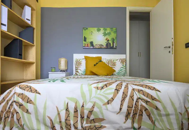 Dormitorio luminoso y bien decorado con colcha estampada, cojines decorativos y una lámpara de noche; estanterías neutras y pared en color de acento completan el ambiente.