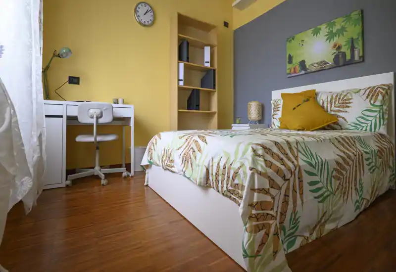 Habitación individual luminosa y bien decorada con colcha estampada, lámpara de mesita y un pequeño escritorio junto a la ventana — ideal para estudiante o persona sola.