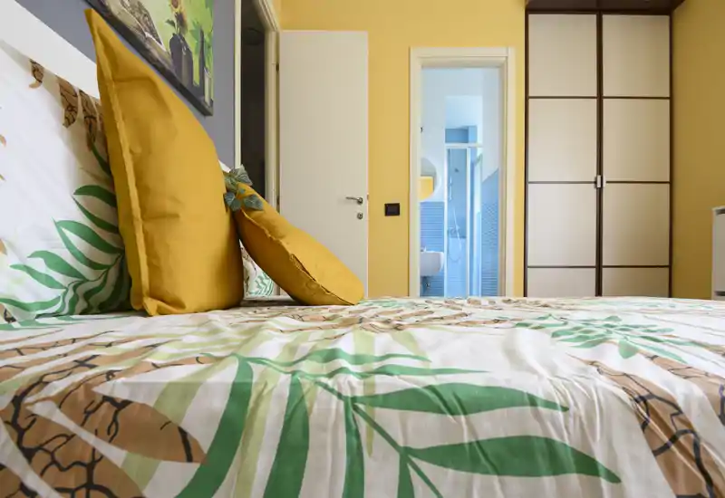 Dormitorio luminoso y decorado con ropa de cama estampada y cojines amarillos; la cama ocupa el primer plano y se ve un baño en suite al fondo.
