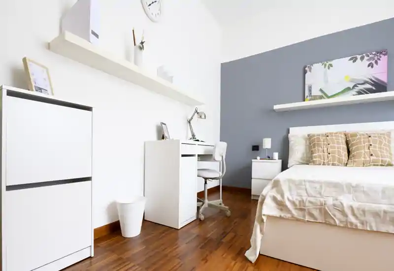 Camera da letto luminosa e minimal con letto matrimoniale ben rifatto, comodino e una scrivania compatta. Pavimento in legno caldo e cuscini di buon gusto creano un'atmosfera moderna e accogliente, perfetta per un annuncio.