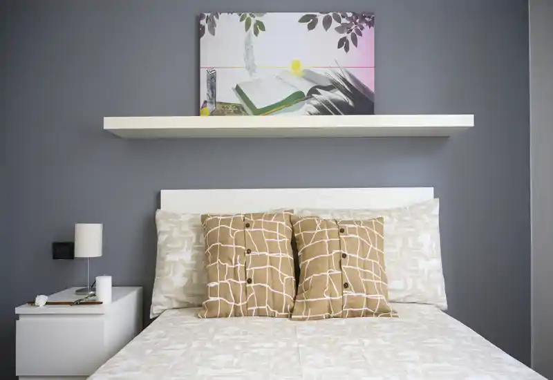 Dormitorio moderno y ordenado con cabecero blanco, cojines con estampado y ropa de cama coordinada, realzado por una balda y un cuadro sobre la cama.