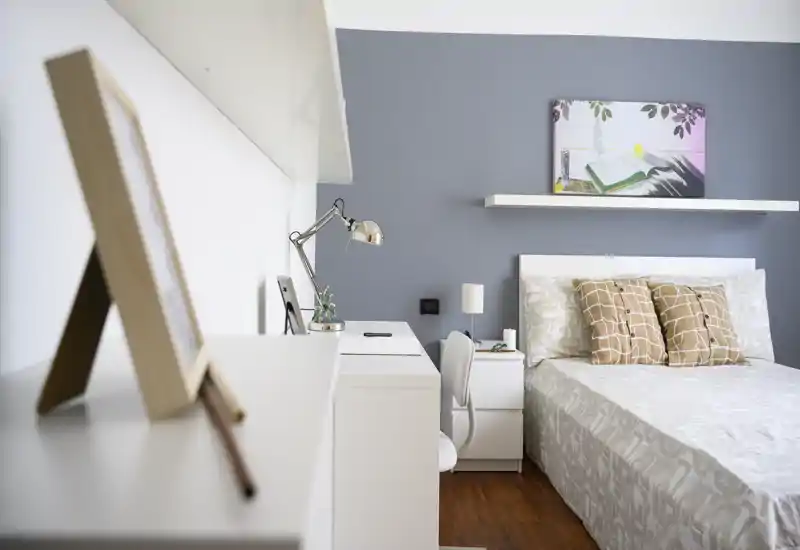 Dormitorio moderno y luminoso con cama doble bien hecha, cojines decorativos, mesita y un pequeño escritorio con lámpara — pared gris y suelo de madera que aportan calidez.