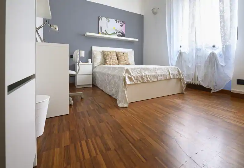 Dormitorio luminoso y ordenado con cama doble, mesita y escritorio; suelo de madera cálida y cortinas translúcidas que aportan una atmósfera acogedora y ligera.