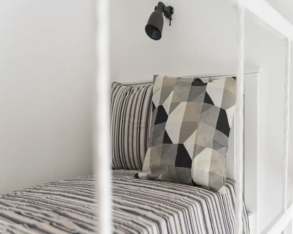 Rincón de dormitorio minimalista y luminoso con ropa de cama a rayas y un cojín estampado sobre cabecero blanco; estilo limpio y aireado con lámpara de lectura en la pared.