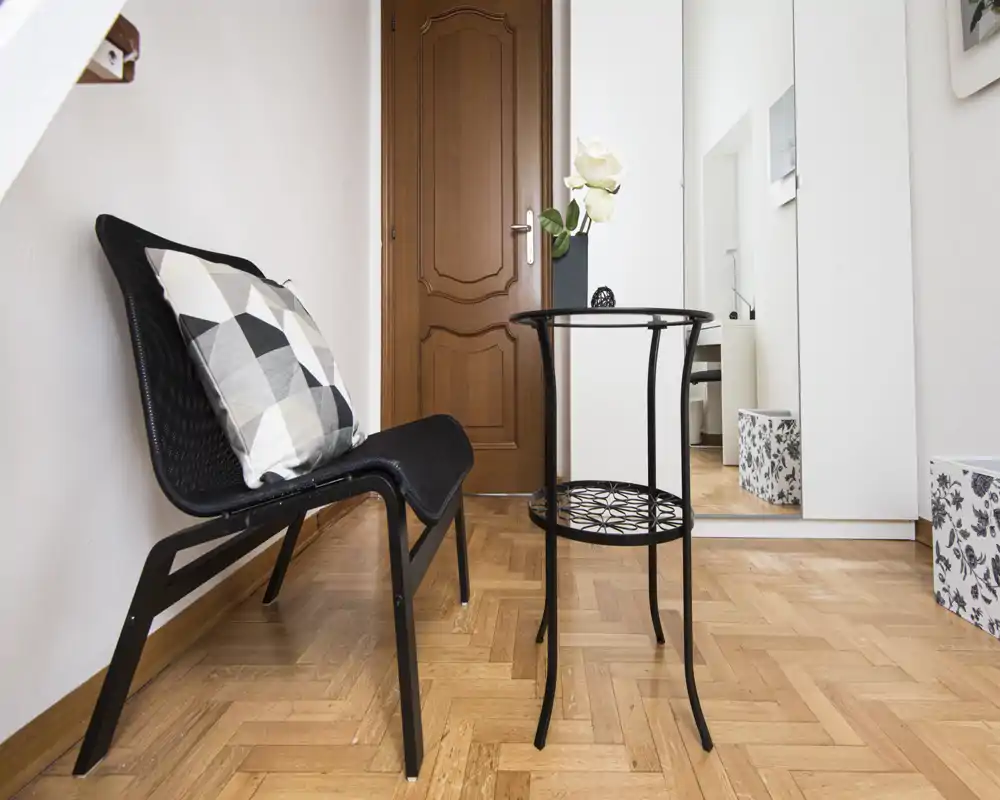 Pequeña entrada bien iluminada con una silla moderna, cojín decorativo y una mesita estrecha; suelo de parquet y espejo de cuerpo entero que dan profundidad.
