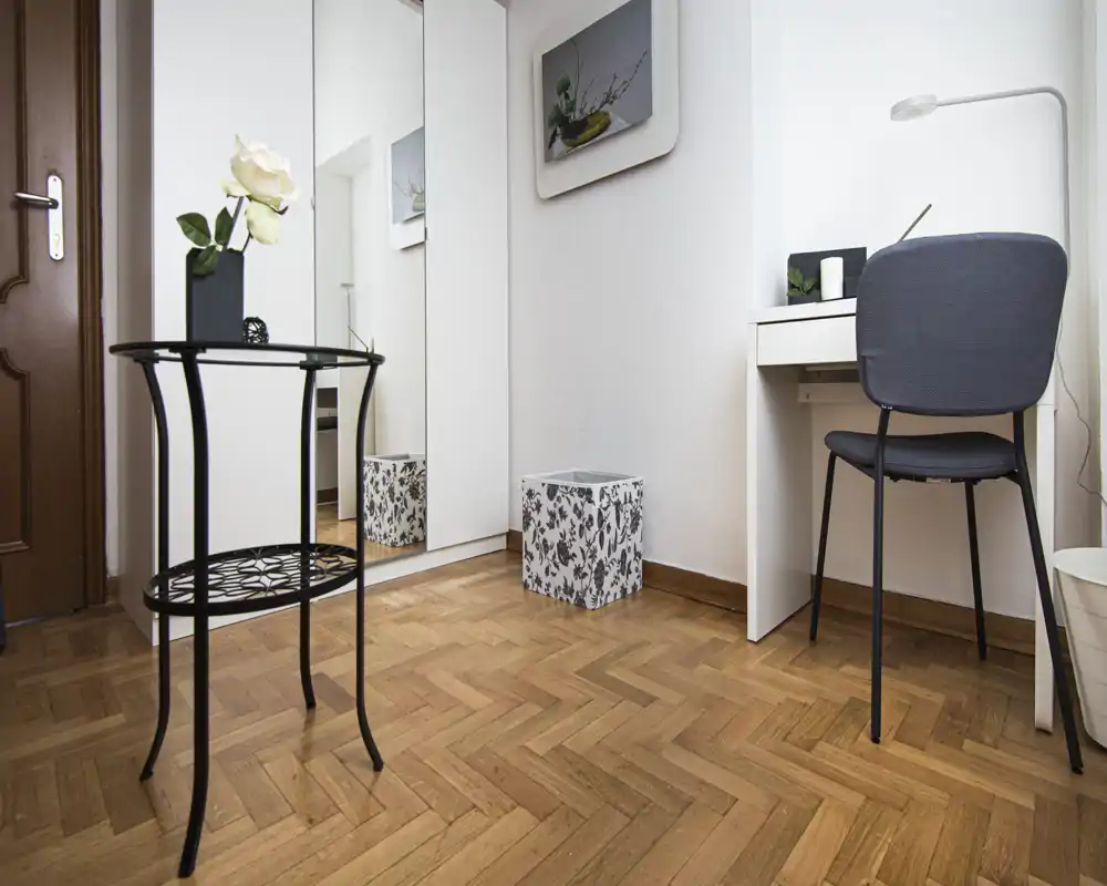 Zona de estar luminosa y de mobiliario sencillo con mesa decorativa, armario con espejo, escritorio y silla — ideal para un estudio o habitación de invitados compacta.