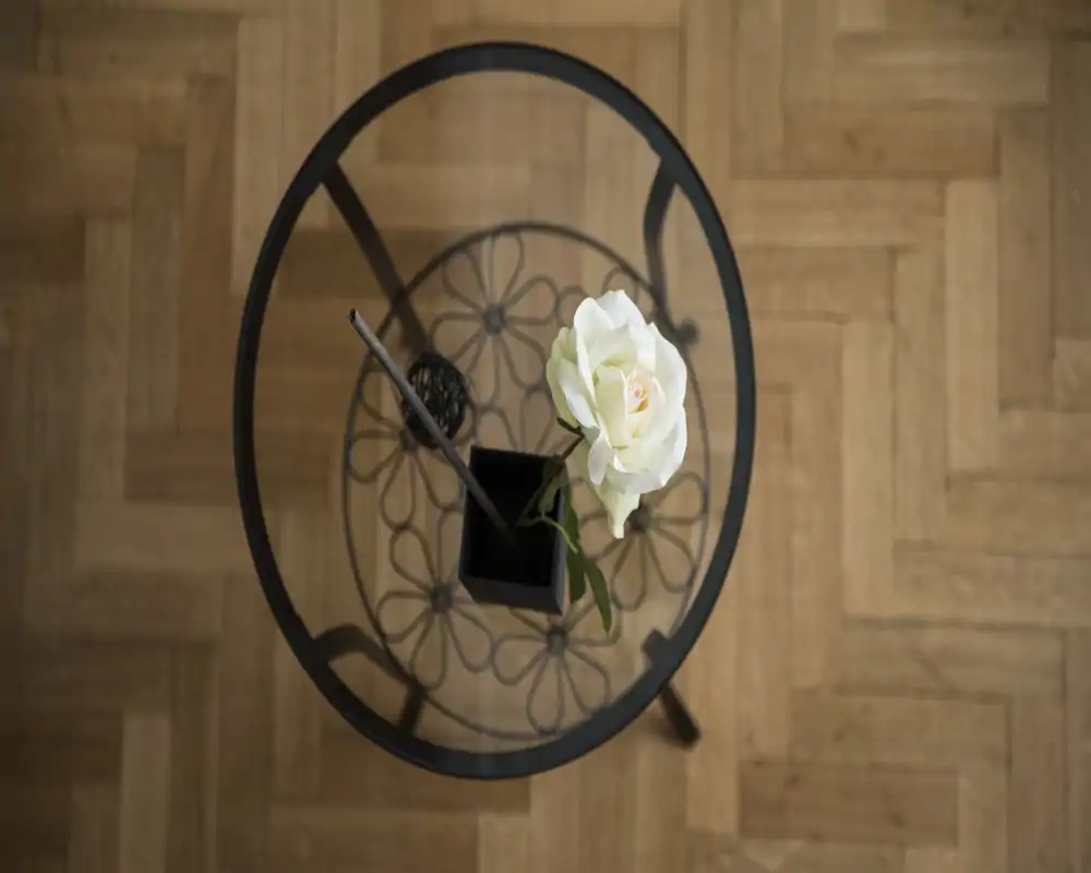 Foto cenital de una pequeña mesa redonda con tapa de cristal y un motivo metálico decorativo, con una rosa blanca en un pequeño jarrón, sobre suelo de parquet.
