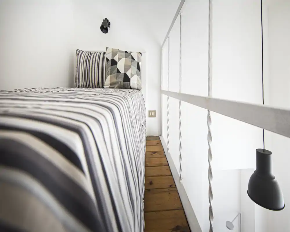 Dormitorio luminoso y minimalista con una cama individual de ropa de rayas y cojines decorativos junto a una barandilla blanca y una lámpara colgante.