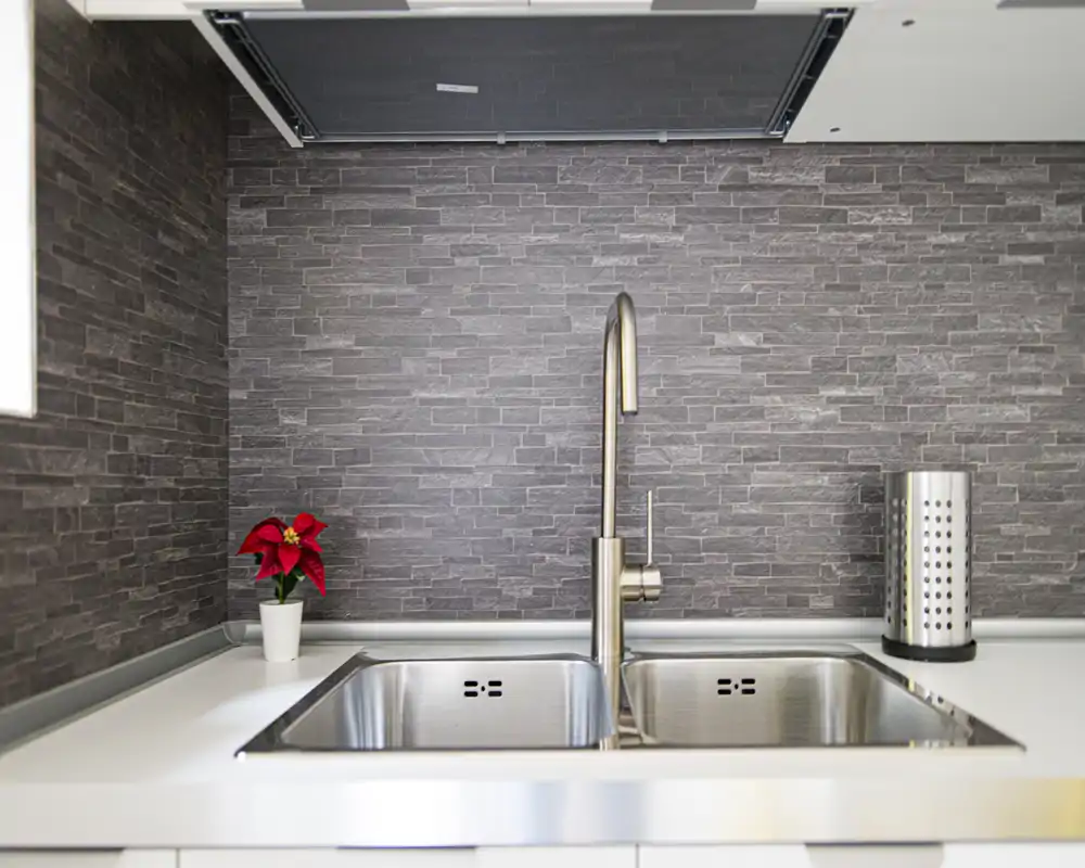 Zona del fregadero de cocina moderna con doble cubeta de acero inoxidable, grifo elegante y salpicadero efecto piedra texturizada. Una planta pequeña y un soporte de utensilios añaden un toque decorativo a la encimera blanca y limpia.