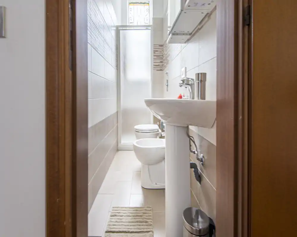 Baño moderno y compacto con lavabo de pie, inodoro y bidé, azulejos en tonos neutros y puerta de ducha con cristal esmerilado; luminoso, limpio y bien aprovechado para espacios pequeños.
