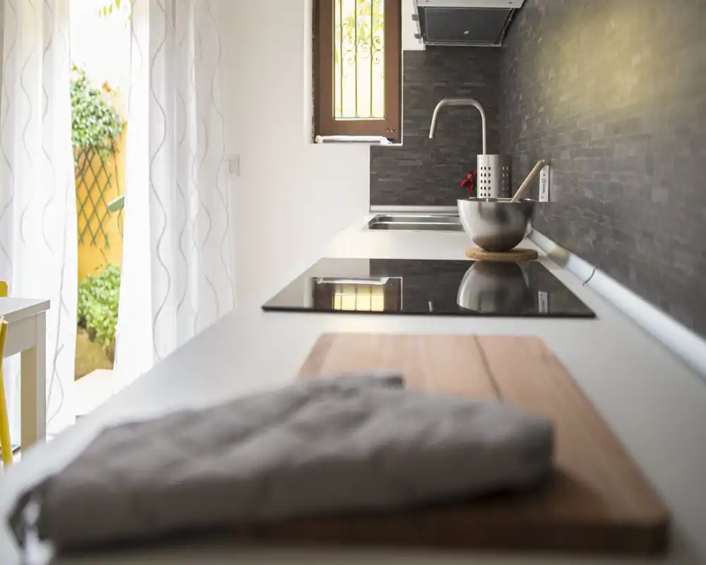 Encimera de cocina moderna y luminosa con placa de inducción, fregadero y accesorios cuidados; cortinas ligeras que se abren a un pequeño espacio exterior aportando luz natural.