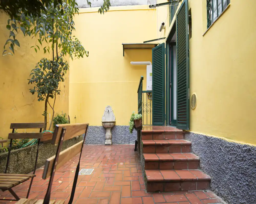 Patio privado soleado con suelos de terracota, asientos exteriores y macetas junto a la entrada del edificio — una pequeña terraza ideal para relajarse.