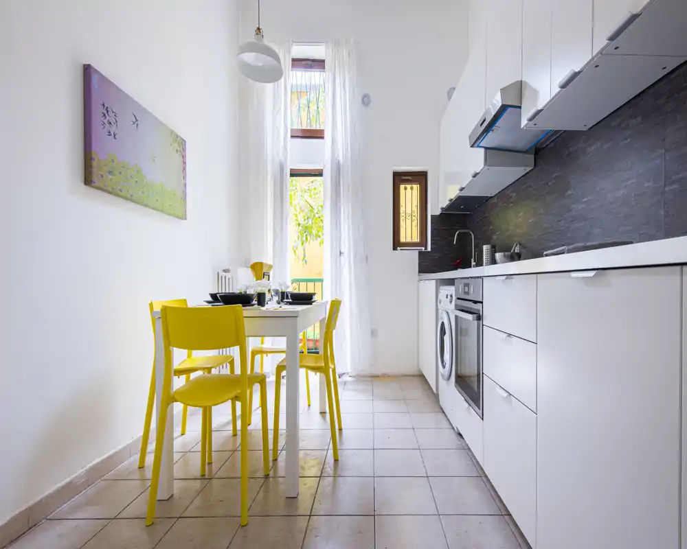 Cocina moderna y luminosa con encimera blanca, electrodomésticos integrados y una mesa pequeña con sillas amarillas alegres; ventana/puerta alta aporta abundante luz natural.