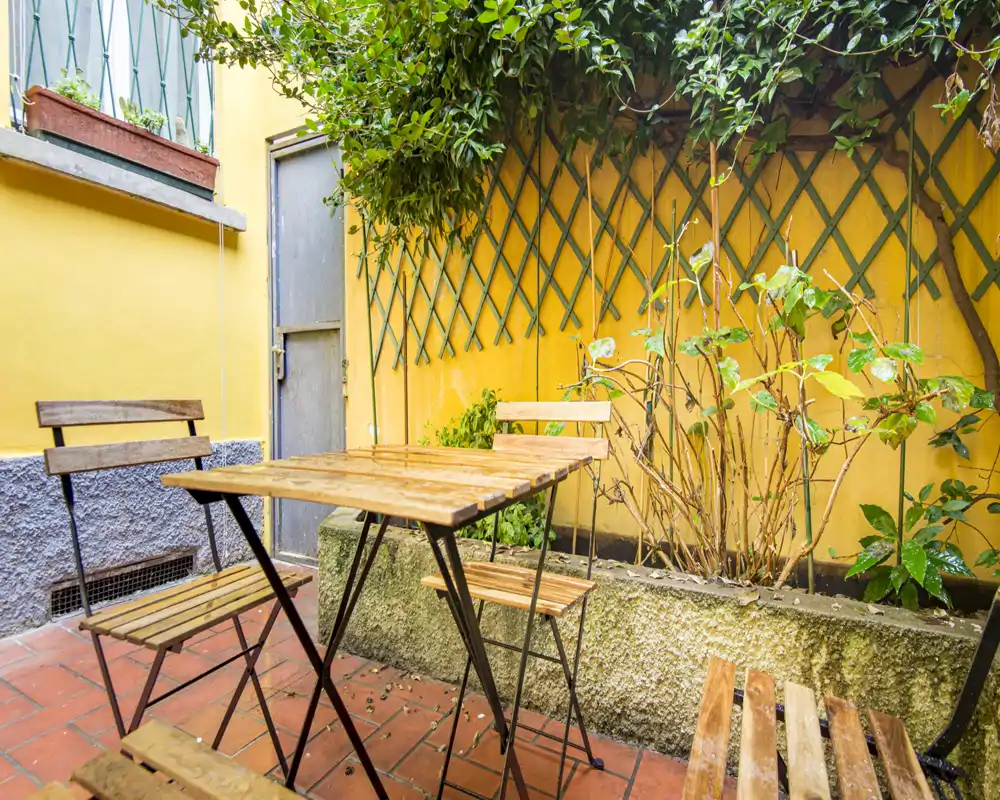 Pequeño patio privado soleado con mesa bistró de madera y sillas plegables, macetas y enrejado sobre una pared amarilla cálida — ideal para el café matutino.