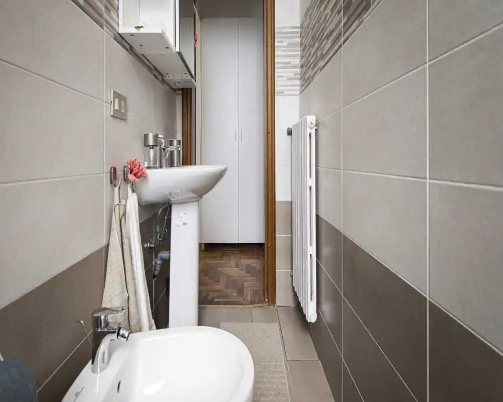 Baño compacto y bien iluminado con lavabo de columna, bidé, ganchos para toallas y radiador; azulejos beige y marrón que ofrecen un aspecto limpio y moderno.