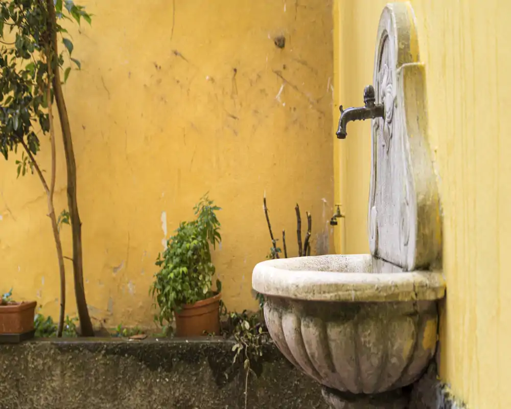 Rincón de un pequeño patio exterior con una pila de piedra decorativa montada en la pared y macetas con plantas frente a una pared amarilla cálida.