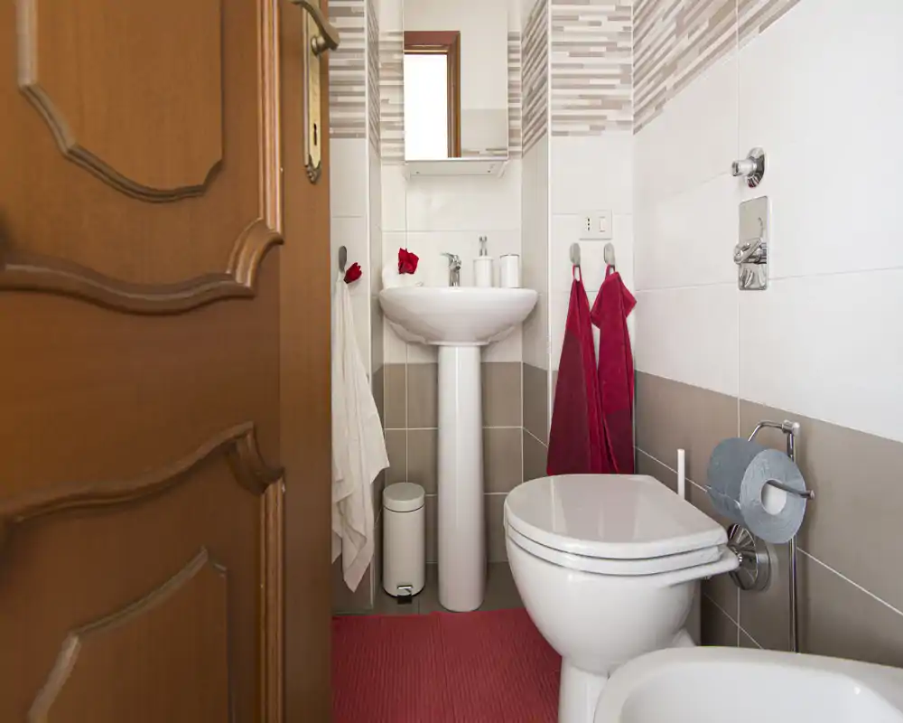 Baño pequeño y cuidado con lavabo de pie, inodoro, bidé y azulejos modernos. Toallas rojas y alfombrilla a juego aportan un toque cálido sobre tonos neutros.